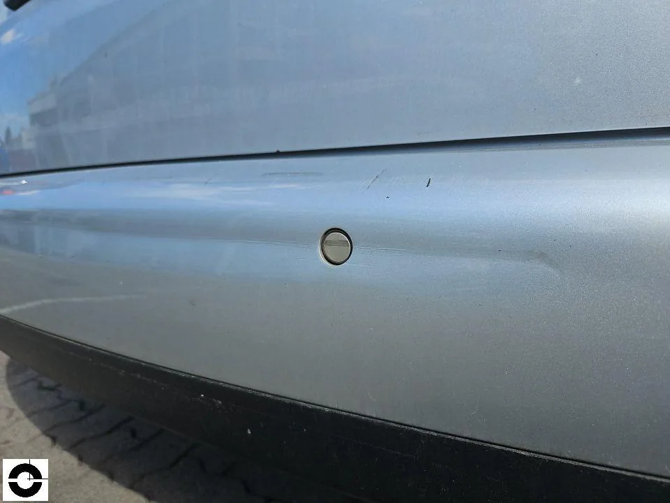 Close-up of a silver rear bumper with a horizontal dent and scratches around a parking sensor