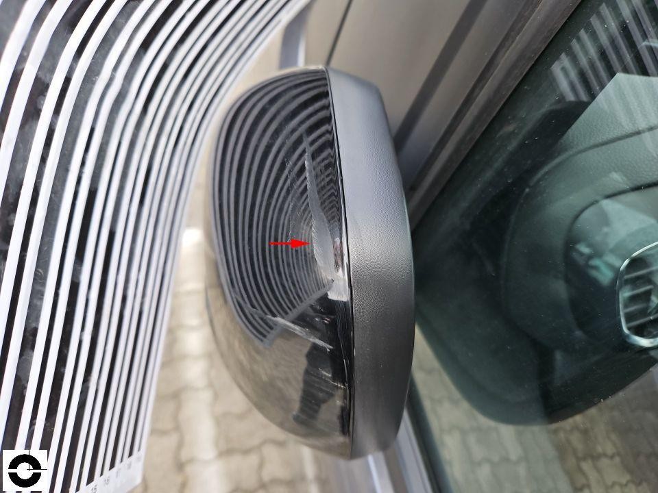 Nahaufnahme linker Außenspiegel mit Beschädigung am Spiegelgehäuse, roter Pfeil markiert Schadstelle