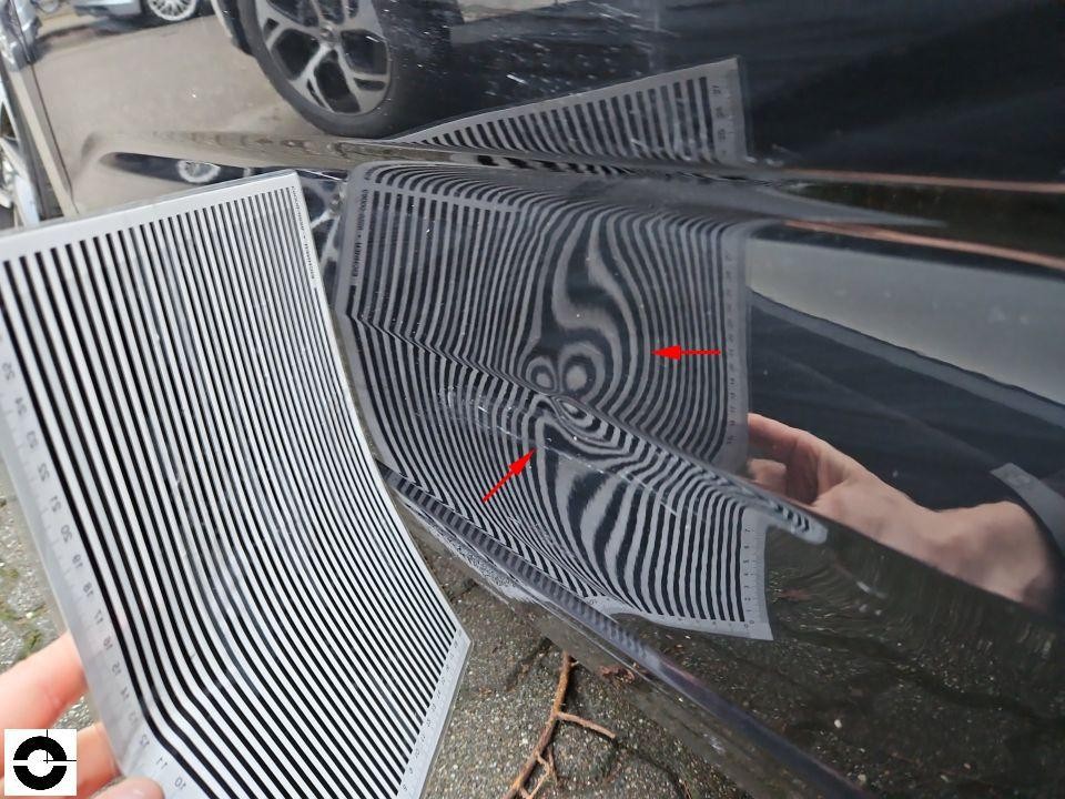 Dellenprüfung mit Streifenlichtbrett an schwarzer Motorhaube, rote Pfeile markieren Dellen