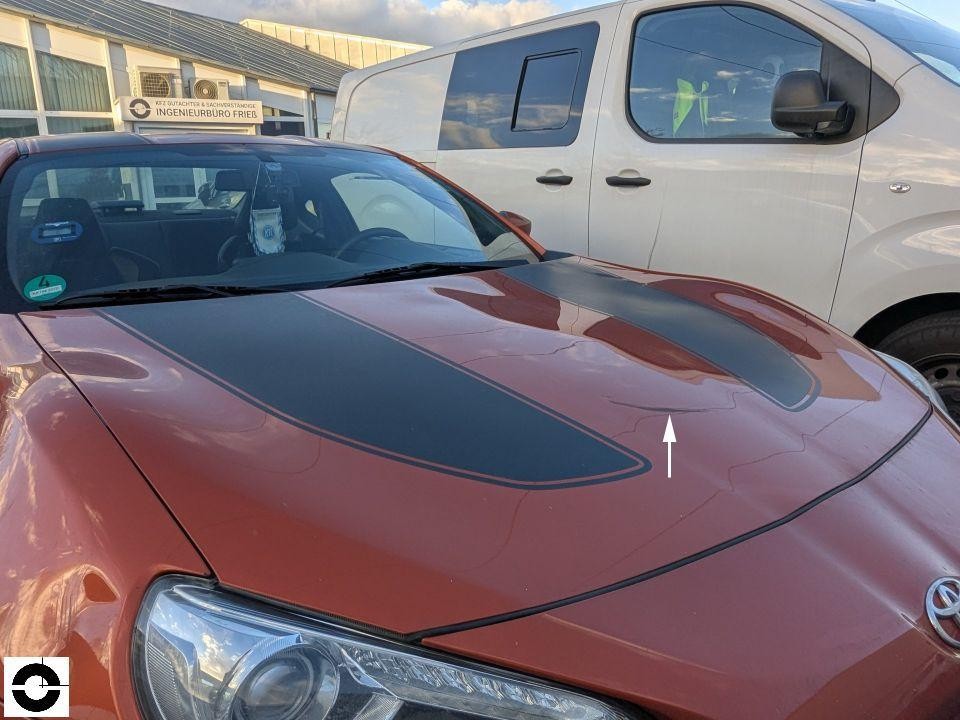 Frontansicht, Motorhaube mit Delle und weißem Pfeil als Schadensmarkierung