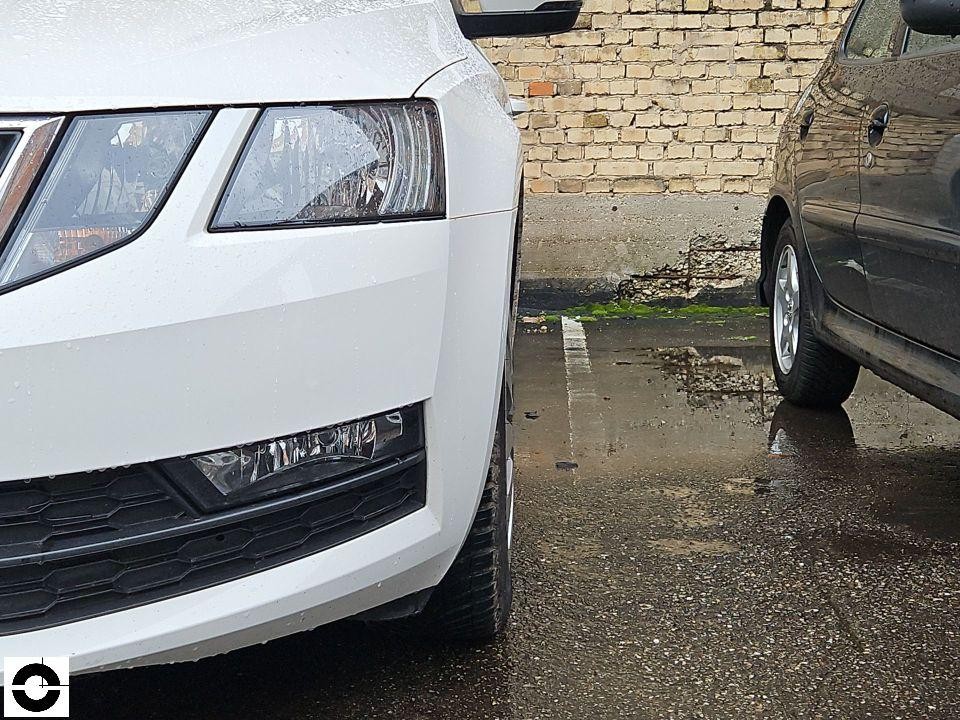 Frontansicht links, weißes Fahrzeug, Stoßfänger und Scheinwerferbereich, nasser Parkplatz